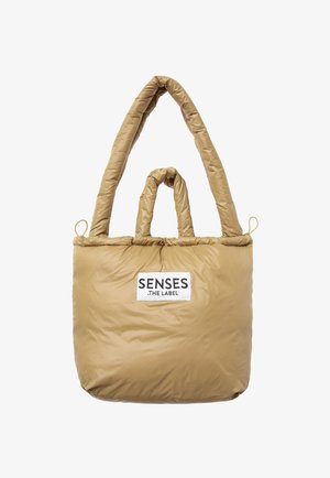 Tan padded tote bag with dual long straps, featuring a rectangular shape and a logo label on the front. Smooth, quilted texture.