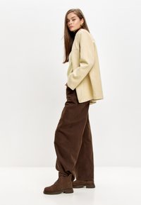 Chaqueta beis de gran tamaño sobre un suéter verde, combinada con pantalones de pierna ancha marrones y botas de cordones marrones. Texturas suaves y estructuradas.