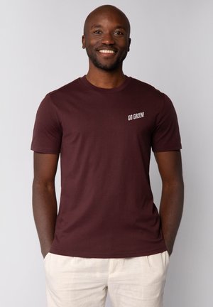 Bordeaux T-shirt van katoen met korte mouwen. Heeft witte tekst "GA GROEN!" op de linkerborst. Gecombineerd met lichtgekleurde broek.