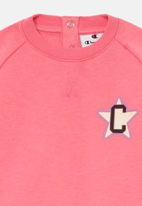 Rózsaszín pulóver, raglán ujjakkal, nagy csillag grafikával és fekete "C" betűvel. Puha pamut anyag, patentos záródással a nyaknál.