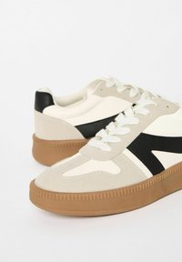 Pair of beige and white sneakers with black detailing and textured brown rubber soles, displayed on a white background.
