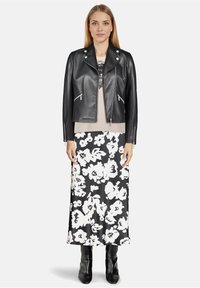 Kobieta w czarnej skórzanej kurtce motocyklowej, beżowym topie, czarno-białej kwiecistej spódnicy i czarnych butach stoi na białym tle.