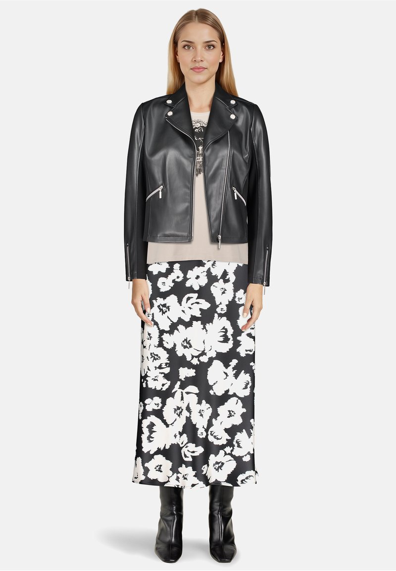 Kobieta w czarnej skórzanej kurtce motocyklowej, beżowym topie, czarno-białej kwiecistej spódnicy i czarnych butach stoi na białym tle.