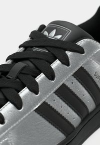 Sudraba krāsas sintētiskas sporta kurpes ar trim melnām svītrām, melnām šņorēm un polsterētu melnu papēdi. Tām ir teksturēts augšdaļa un reljefa logotipa detaļa.