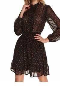 Vestido negro hecho de tela transparente, con un patrón floral con corazones rojos, mangas largas abullonadas, cintura fruncida y un dobladillo con volantes.