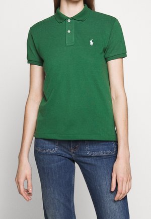 Vrouw draagt een groen poloshirt met korte mouwen en een wit geborduurd logo, gecombineerd met blauwe spijkerbroeken.