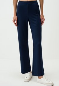 Marineblauwe flared broek van zachte, rekbare stof met een gladde textuur en een tailleband. Draagt comfortabel met witte sneakers voor een contrasterende look.