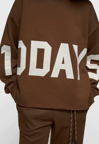 Sweat-shirt marron surdimensionné présentant un grand graphisme blanc "10 DAYS" sur le devant. Tissu doux avec une coupe décontractée. Pantalon assorti marron avec cordon de serrage.