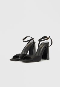 Μαύρα σαντιγί πέδιλα με ψηλό τακούνι, ανοιχτή μύτη, λουράκι στον αστράγαλο και τετράγωνο τακούνι. Διαθέτουν γυαλιστερή μαύρη λεπτομέρεια στο τακούνι.