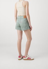 Ljust gröna denimshorts med bakfickor och en märkeslogotyp i läder. Modellen bär en beige linne och krämfärgade sneakers med röda ränder.