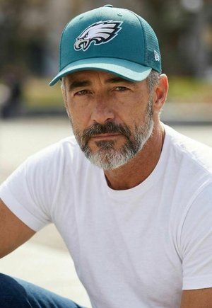 Homme avec barbe poivre et sel portant une casquette de baseball bleu sarcelle avec un logo d'aigle et un t-shirt blanc, en extérieur avec un arrière-plan flou.