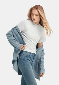 T-shirt bianca a maniche corte sotto una giacca di jeans azzurri chiari e jeans blu. La giacca ha bottoni metallici e una vestibilità comoda.