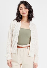 Veste bomber en dentelle crème avec des poignets et un col côtelés. Portée sur un débardeur vert olive, associée à un pantalon beige et une ceinture marron.