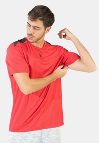 Camiseta deportiva roja de manga corta con acentos negros en los hombros y un logo en el pecho. Ligera, de textura suave, adecuada para ropa activa.