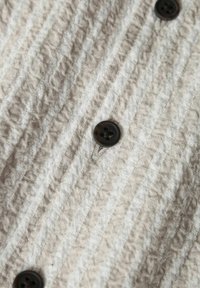 Ljust beige textil med strukturerad yta som har vertikala ränder och en mjuk, skrynklig finish. En mörkbrun knapp är synlig.