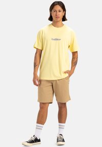 Camiseta de algodón amarilla clara con logo, combinada con pantalones cortos color beige. El modelo lleva zapatillas negras y calcetines blancos con rayas grises. Ajuste casual.