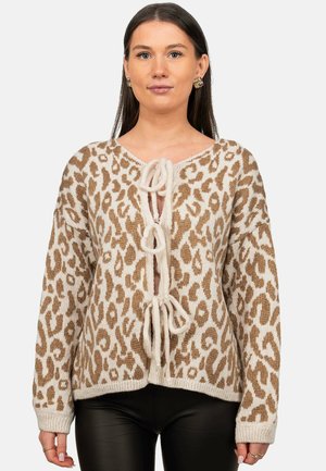 Frau trägt eine beige-braune Strickjacke mit Leopardenmuster und drei vorderen Bindeschließen sowie schwarze Hose und schaut nach vorne.