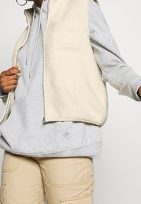 Beżowa kamizelka z polaru z wysokim kołnierzem, pojedynczą kieszenią na piersi i zapięciem na zamek. Nałożona na szary polarowy sweter z kapturem. Widoczne beżowe spodnie cargo.