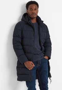 Man wearing a navy quilted long jacket with a hood, dark blue henley shirt, and blue jeans against a white background.