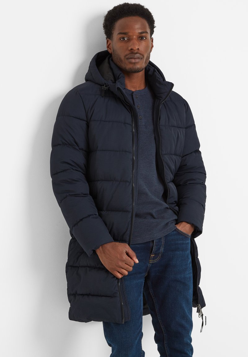 Man wearing a navy quilted long jacket with a hood, dark blue henley shirt, and blue jeans against a white background.