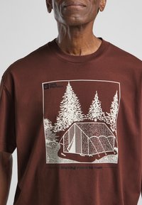 Bruine katoenen T-shirt met een witte gedrukte graphic van een tent en bomen. De tekst eronder leest "Making room to roam." Rechthoekig ontwerp, korte mouwen.
