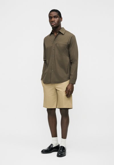 Homme debout portant une chemise boutonnée olive à manches longues, un short beige jusqu'aux genoux, des chaussettes blanches et des mocassins noirs sur un fond uni.