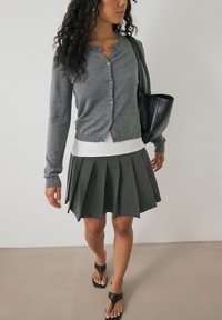 Cardigan grigio a bottoni, canotta bianca a coste, gonna grigia plissettata e sandali neri. Gli accessori includono una borsa a tracolla nera.