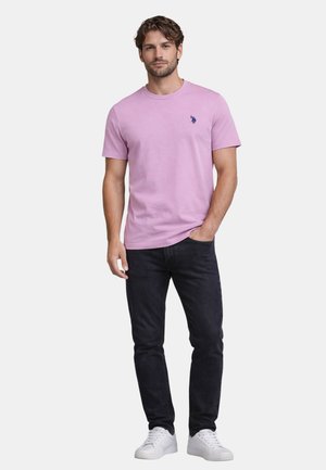 Mann mit kurzen lockigen Haaren, der ein hellviolettes kurzärmeliges Hemd, schwarze Jeans und weiße Turnschuhe trägt und mit einer Hand in der Tasche steht.