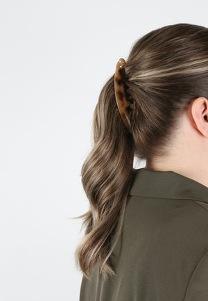 Schildpatt-Haarspange, die welliges braunes Haar zu einem Pferdeschwanz zusammenhält. Sie hat ein geschwungenes Design mit Zähnen für besseren Halt und ist vor einem einfarbigen Hintergrund platziert.