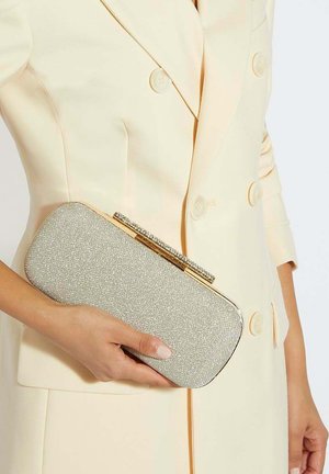 Woman in cream double-breasted blazer holding a silver glittery rectangular clutch with a jeweled clasp.