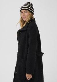 Cappotto nero strutturato con colletto, dettaglio di grandi bottoni e tasche laterali. Abbinato a un berretto a strisce nero e crema.