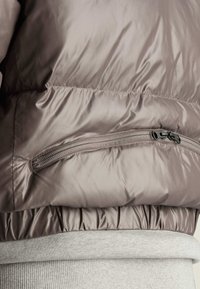 Padded jacket w kolorze jasnego taupe, z gładkim, błyszczącym wykończeniem, elastycznym paskiem w talii oraz zapinaną na zamek kieszenią z boku.