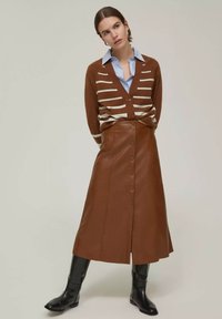 Cardigan a righe marroni sopra una camicia blu, abbinato a una gonna A-line in pelle marrone con bottoni frontali e stivali neri al ginocchio.