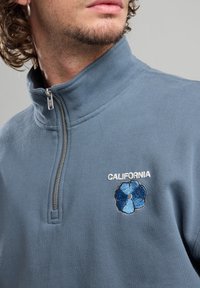 Light blue fleece sweater with a high collar, half-zip closure, and embroidered blue flower design; "CALIFORNIA" text in white.