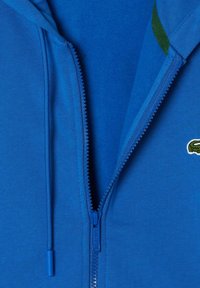 Sweat-shirt zippé bleu avec une texture lisse, présentant une fermeture éclair sur le devant et un petit logo brodé vert sur la zone gauche de la poitrine.