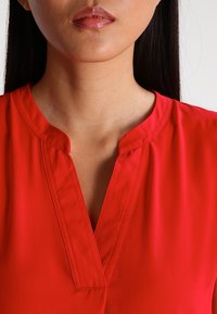 Blouse rouge légère avec un décolleté en V et des plis subtils. Texture lisse avec des manches courtes, conçue pour une coupe ajustée.