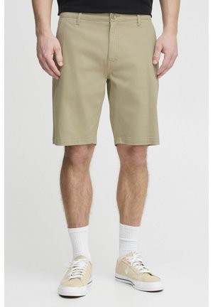 Homme portant un short beige mi-genoux, des chaussettes blanches et des baskets beiges, debout devant un fond uni.