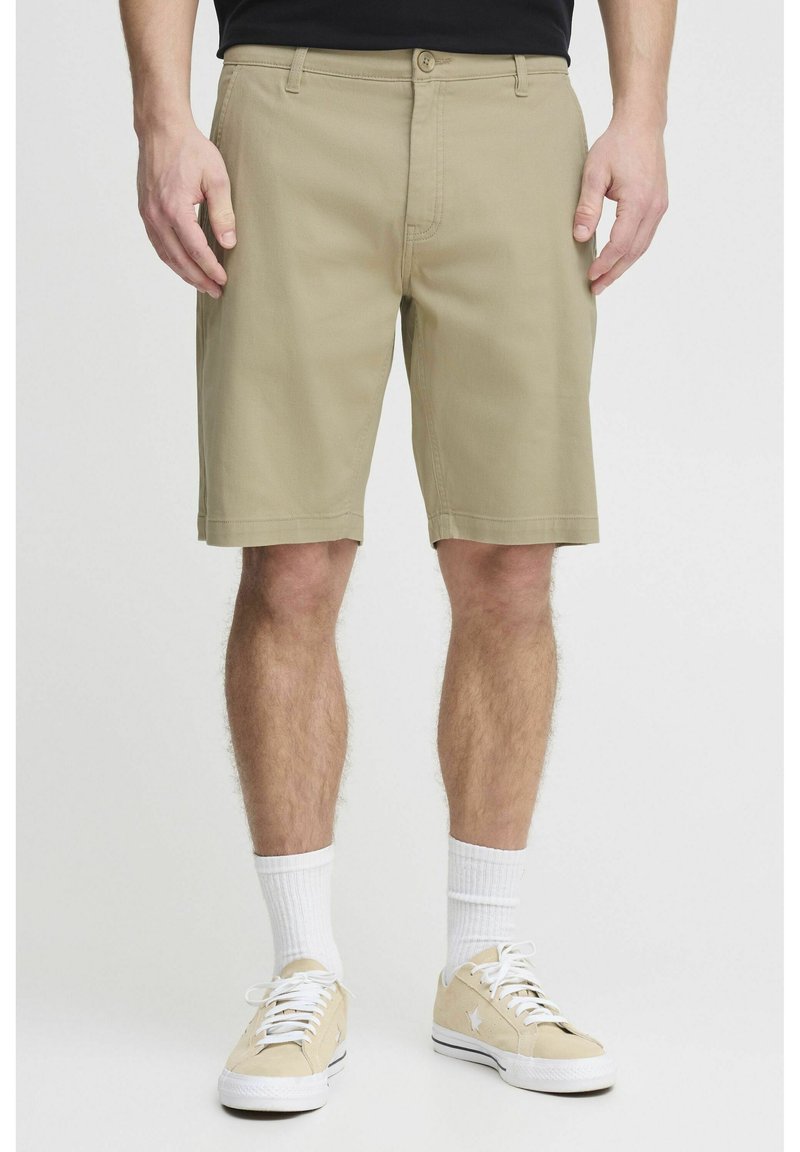 Homme portant un short beige mi-genoux, des chaussettes blanches et des baskets beiges, debout devant un fond uni.