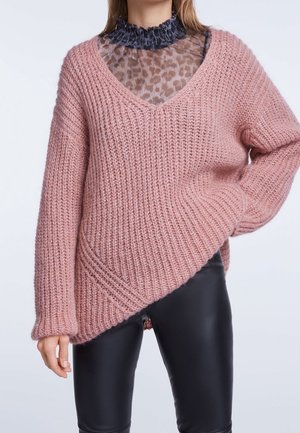 Vrouw draagt een roze grove gebreide V-hals trui over een transparante top met luipaardprint, gerimpelde kraag en zwarte nep leren broek.