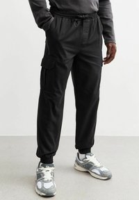Zwarte cargobroek met elastieke tailleband, zijzakken en boorden. Gecombineerd met grijze sneakers met een gestructureerd patroon en witte accenten.