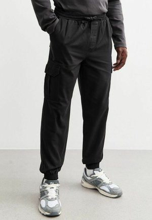 Pantalons cargo noirs avec taille élastique, poches latérales et poignets. Portés avec des baskets grises présentant un motif texturé et des accents blancs.