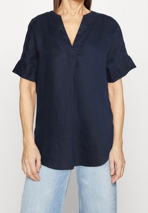 Vrouw draagt een marineblauwe blouse met korte mouwen, V-hals en opgerolde mouwen, gecombineerd met lichtblauwe spijkerbroek, staand voor een effen achtergrond.