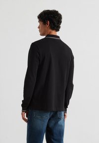Schwarzes Langarm-Polohemd mit flachem Kragen, ausgestattet mit weißen Akzenten am Hals und einem geraden Saum, kombiniert mit blauen Jeans.