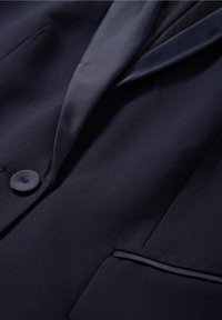 Laivansinis bleiseri, joka on valmistettu sileästä kankaasta, sisältää notch-lapelsin, yhden napin ja hienovaraisen taskudetaillin.