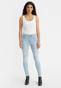Mujer con cabello largo y oscuro, vestida con una camiseta sin mangas blanca, jeans ajustados rasgados de color azul claro y botas negras hasta el tobillo, de pie contra un fondo blanco liso.