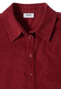 Chemise en velours côtelé rouge avec col et trois boutons sur le devant, avec une étiquette portant le nom de la marque Cecil.