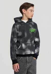Ragazzo con capelli castani che indossa un hoodie a motivi tie-dye grigio scuro e bianco con un grafico di un controller di gioco verde neon e maniche nere risvoltate, con le mani in tasca.