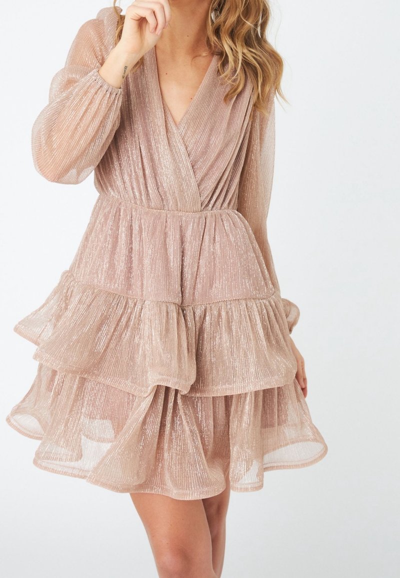 Femme portant une mini-robe à manches longues nude-rose brillante, superposée, avec un col en V et un détail de tissu plissé.