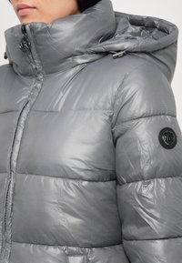 Personne portant une doudoune grise brillante avec un col montant, une capuche, et un patch logo circulaire noir visible sur la manche.