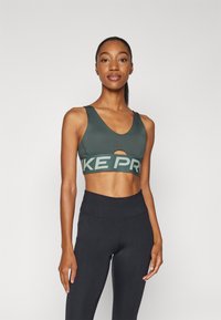 Športni modrček v temno zeleni barvi z V-izrezom in izrezom. Ima širok pas s svetlo sivo napisom "NIKE PRO". V kombinaciji s črnimi leggingsi z visokim pasom.